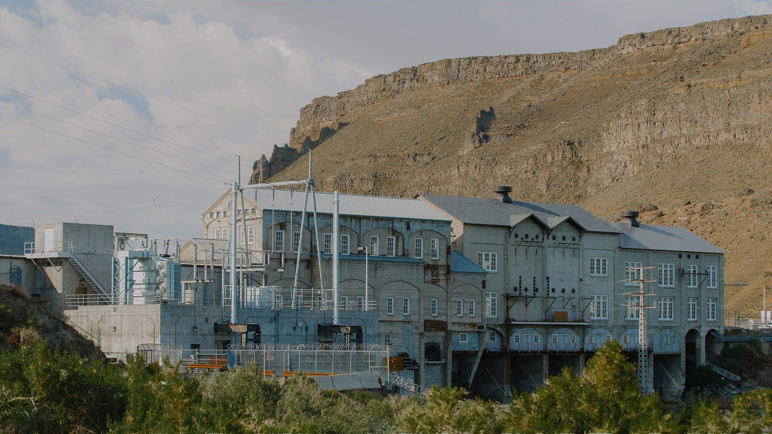 a building attached to a dam with treetops in the foreground and mountains in